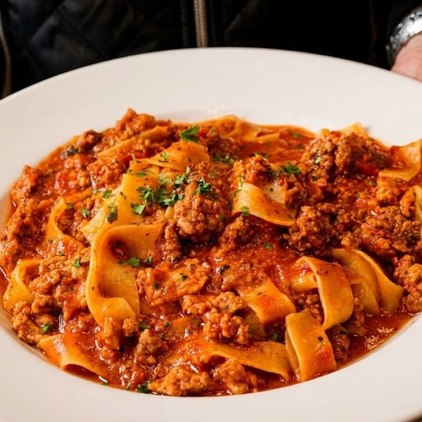 Hands holding a white plate filled with pappardelle pasta and a rich meat ragu, garnished with parsley.