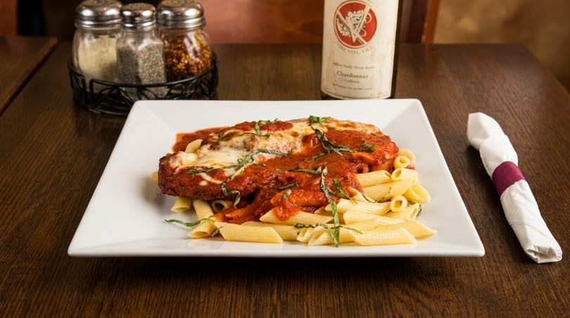 Chicken parmesan with penne pasta, tomato sauce, and basil, alongside a wine bottle and spices.