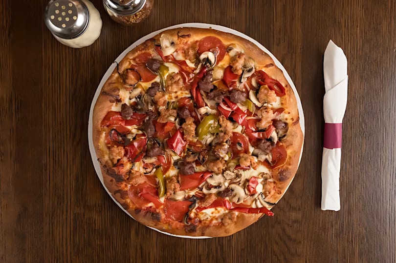 Overhead view of a supreme pizza with pepperoni, sausage, peppers, and mushrooms on a wooden table.